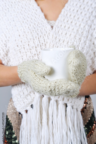 Woman wearing knitted scarf and mittens holding white cup of warming drink in her hands closeup
