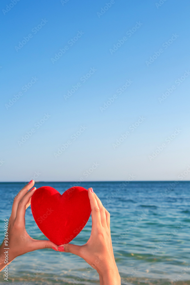 Fototapeta premium Hands holding red heart with blue sky and sea horizon in the background.