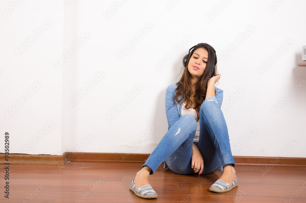 Pretty brunette wearing denim jeans and shirt plus white top sitting on wooden surface her back against wall, black headphones listening to music Pretty brunette wearing denim jeans and shirt plus white top sitting on wooden surface her back against wall, black headphones listening to music