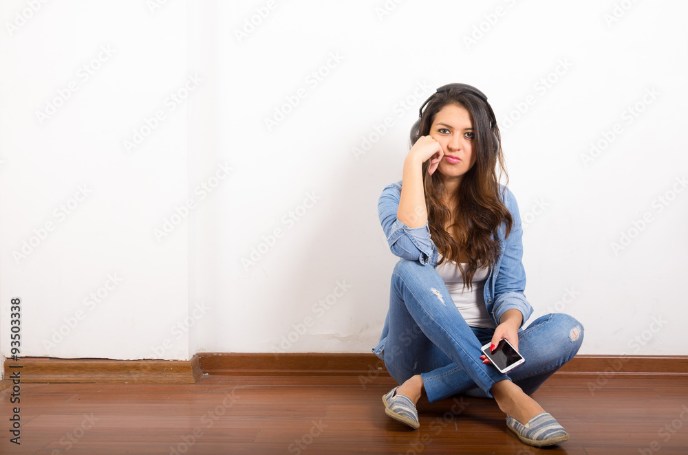 Pretty brunette wearing denim jeans and shirt plus white top sitting on wooden surface her back against wall, black headphones listening to music Pretty brunette wearing denim jeans and shirt plus white top sitting on wooden surface her back against wall, black headphones listening to music