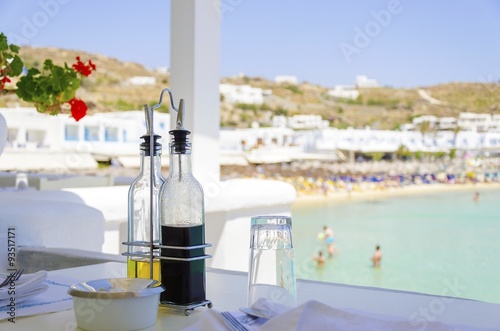 A table set with plates, glasses, olive oil and balsamic vinegar on a beach bar restaurant and a view of the blue sea in Mykonos, Greece. A greek island scene at the beach ready for lunch.