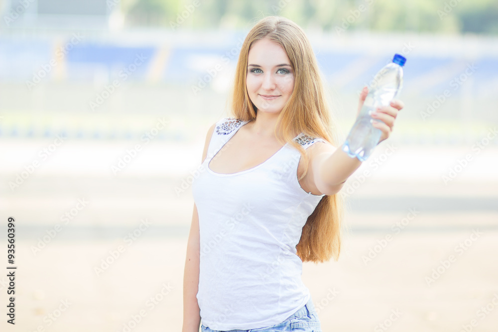 Selective focus on pretty young blond-haired woman wearing white T-shirt standing aside drinking water and smiling satisfied with her life. Park on background Selective focus on pretty young blond-haired woman wearing white T-shirt standing aside drinking water and smiling satisfied with her life. Park on background