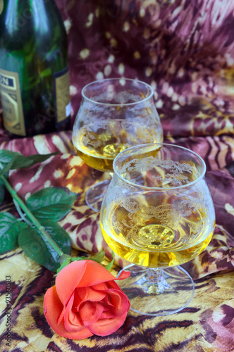 Two glasses of cognac with red roses and a bottle on a colored background
