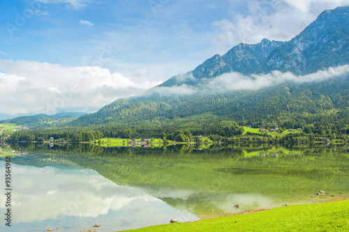 Coast covered with green grass, beautiful mountains, white clouds, blue sky and picturesque houses with reflection in the lake.