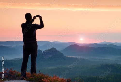 Man takes photos with smart phone on peak of rock empire. Dreamy fogy landscape, spring orange pink misty sunrise above beautiful valley