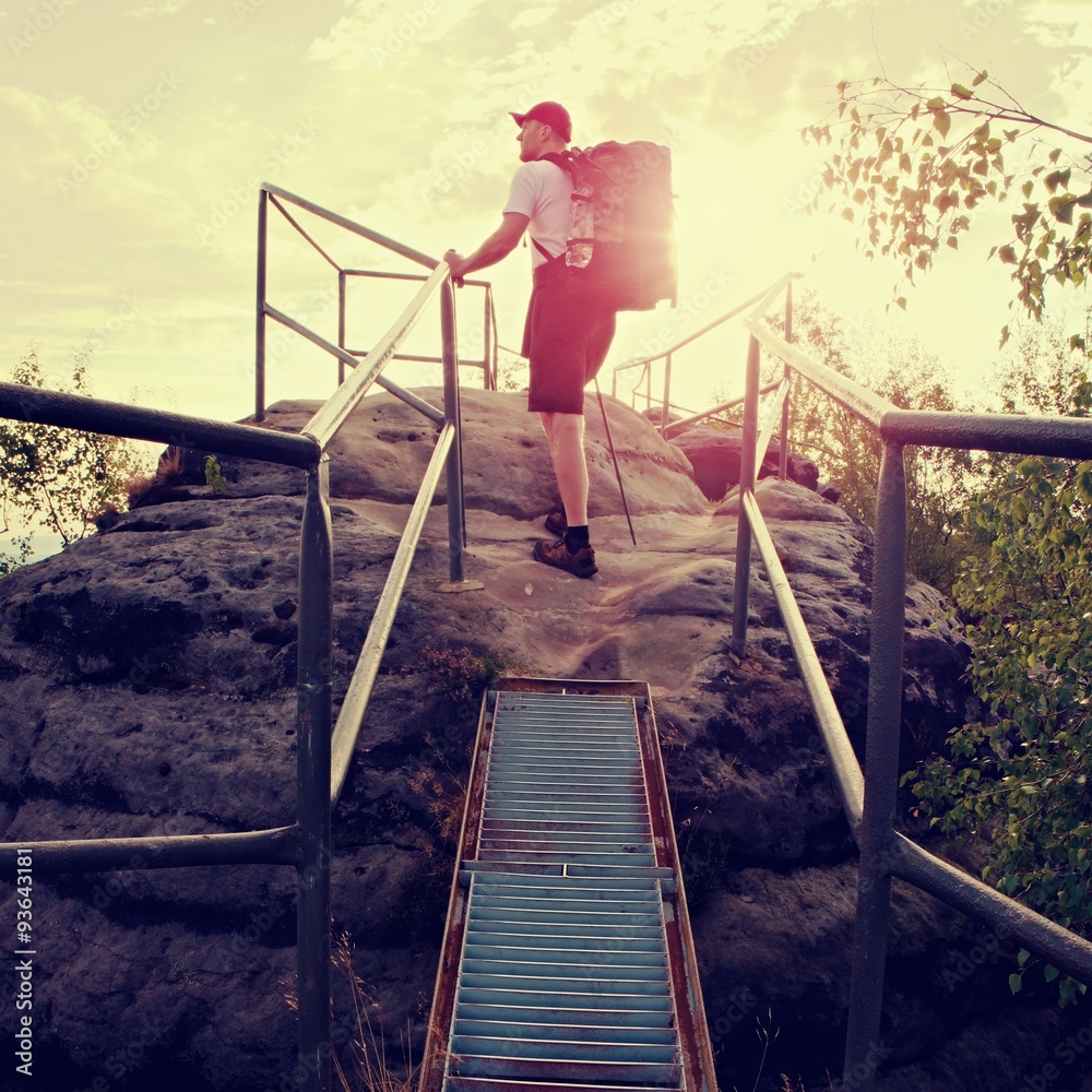 Tall backpacker hold handrail on rock. Sunny daybreak in rocks. Hiker with big backpack, baseball cap, dark pants and white shirt. Tall backpacker hold handrail on rock. Sunny daybreak in rocks. Hiker with big backpack, baseball cap, dark pants and white shirt.