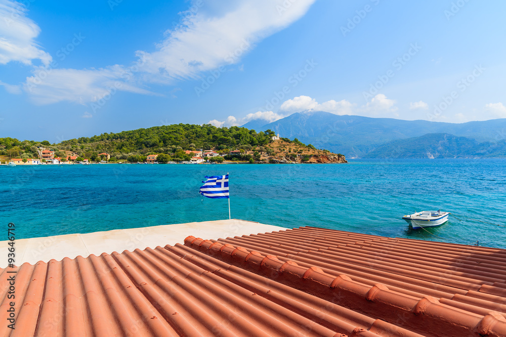 Obraz premium Orange tile roof of a house with Greek flag waving against blue sea background on coast of Samos island, Greece