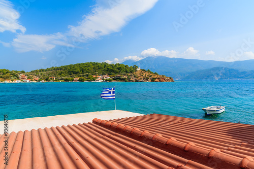 Orange tile roof of a house with Greek flag waving against blue sea background on coast of Samos island, Greece