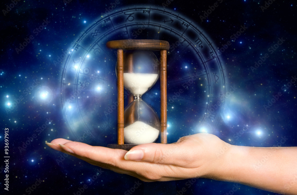 female hand holding an hourglass over blue mystical background with stars and astrology chart - obrazy, fototapety, plakaty 