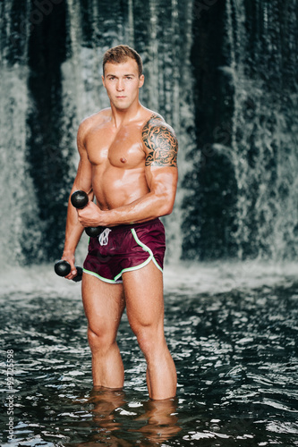 Muscular man lifting dumbbell on waterfall