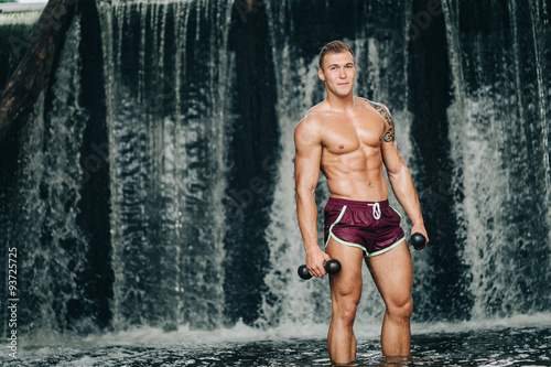 Muscular man lifting dumbbell on waterfall