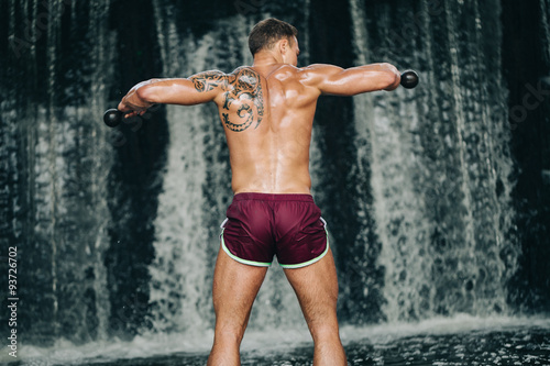 Muscular young man lifting dumbbell on waterfall