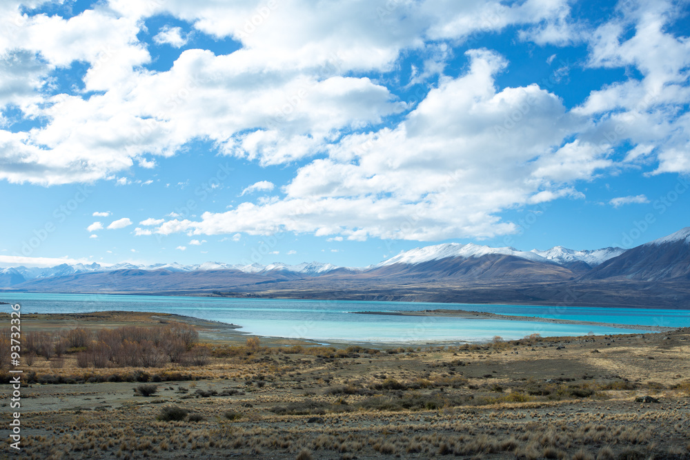 Lake Tekapo in beautiful turquoise blue, that is caused by glacial flour, fine rock particles from the glaciers. The lake is dominant among brown dry grass, and dark-toned color of mountain ridges.
