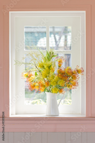 Artificial flowers in white vase beside white window.