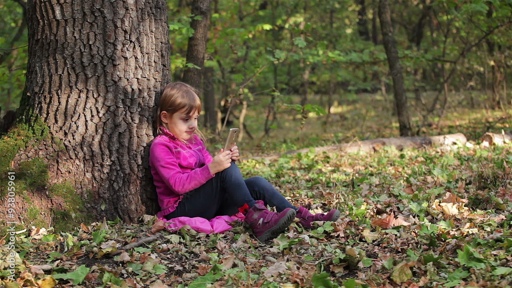 Little girl playing on a smartphone in the forest, sitting under a tree Little girl playing on a smartphone in the forest, sitting under a tree