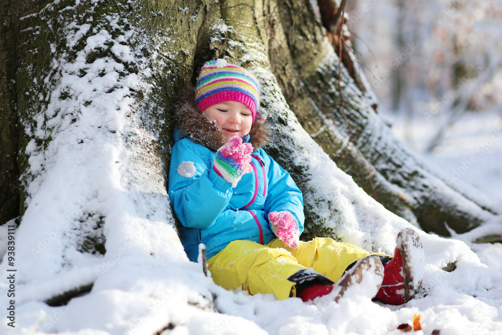 Active healthy toddler girl wearing colorful snowsuit and bright knitted hat and mittens enjoying snow playing in a beautiful snowy forest on sunny winter day Active healthy toddler girl wearing colorful snowsuit and bright knitted hat and mittens enjoying snow playing in a beautiful snowy forest on sunny winter day