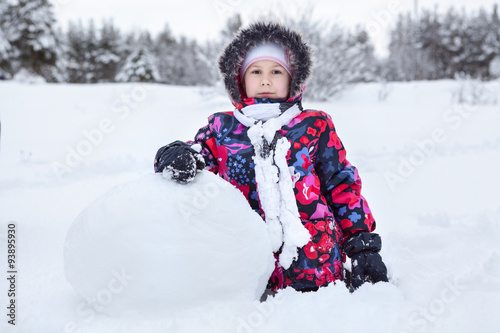 Pretty Caucaian girl in winter clothes rolling big snowball for snowman making, winter
