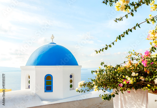 Blue dome of church on Santorini island in the early morning with tropical flower plant and yellow sunlight, Mediterranean sea, Greece
