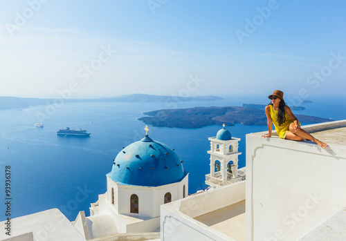 Female tourist in yellow dress, wearing hat enjoying view on Santorini in the morning on building above famous church, Mediterranean sea, Greece