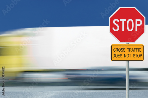 Red stop road sign, motion blurred truck vehicle traffic in background, regulatory warning signage octagon, white octagonal frame, metallic pole post, yellow cross traffic does not stop text signage