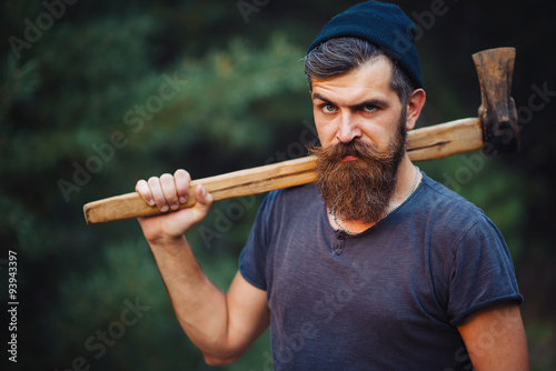 Brutal brunette bearded man in warm hat with a hatchet in the woods on a background of trees photo
