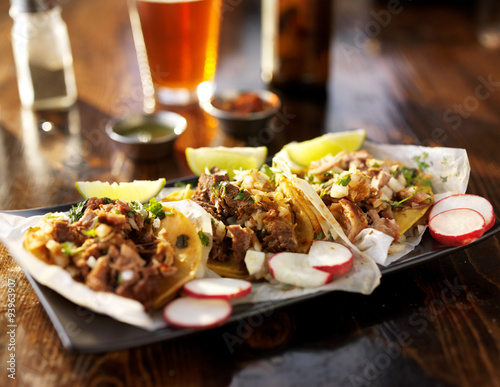 three tacos with beer on wooden table top served with limes and radishes