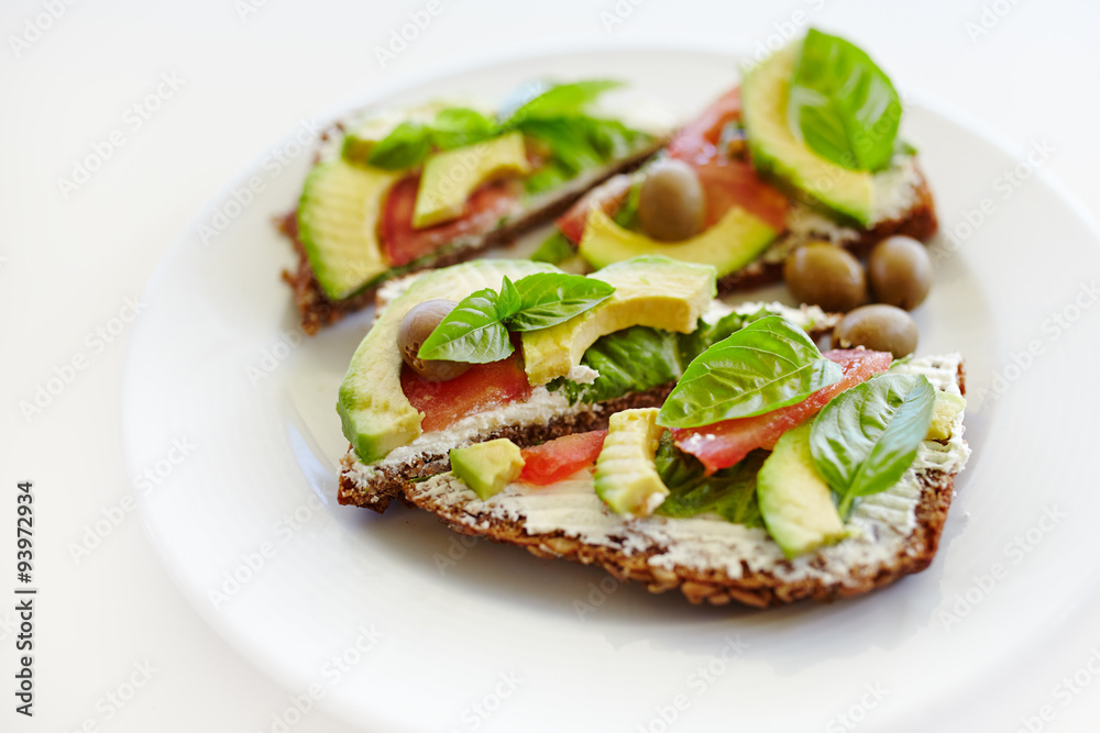 Healthy sandwich with whole-grain bread, avocado, tomato, basil leaves and olives on plate; healthy eating; organic food;
