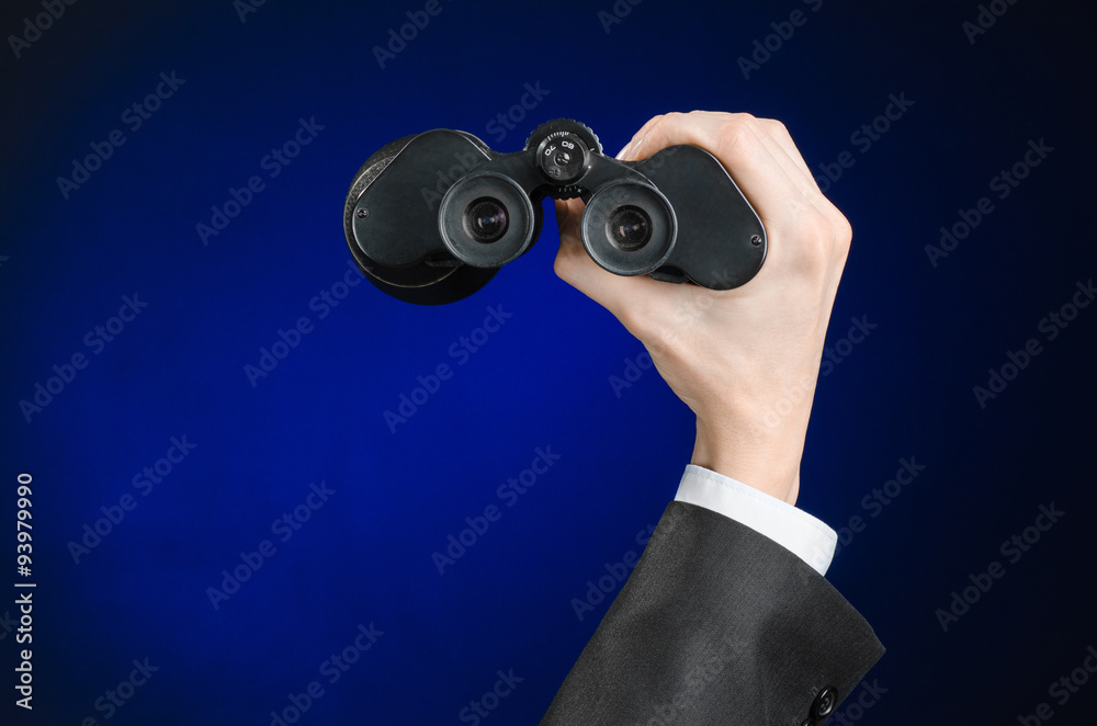 Business and search topic: Man in black suit holding a black binoculars in hand on a dark blue background in studio isolated Business and search topic: Man in black suit holding a black binoculars in hand on a dark blue background in studio isolated