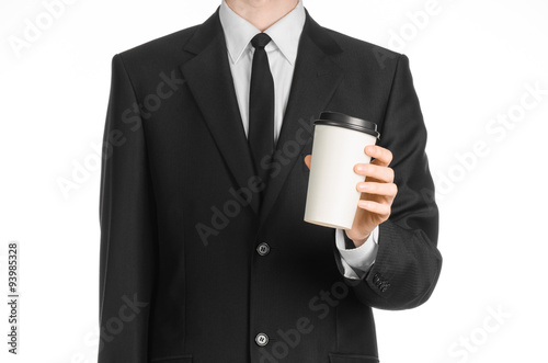 Business lunches coffee theme: businessman in a black suit holding a white blank paper cup of coffee with a brown plastic cap isolated on a white background in the studio, advertising coffee