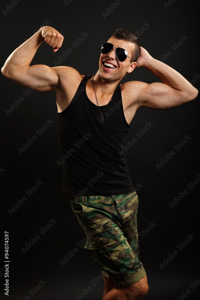 Young smiling military man funny poses and shows his biceps on dark background Young smiling military man funny poses and shows his biceps on dark background