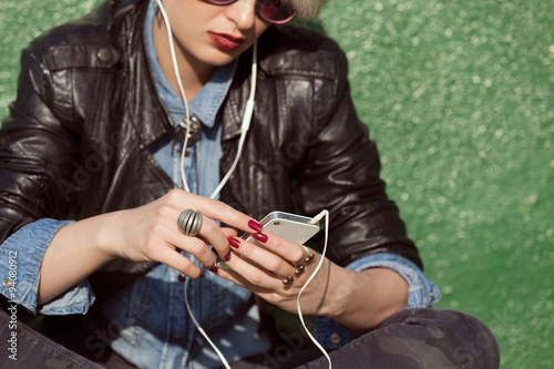 young blond woman with punk style hair using her mobile phone and leaning on green wall
