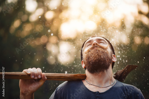 Tableau sur toile bearded brutal man in warm hat lumberjack with an ax in a forest at sunset, and