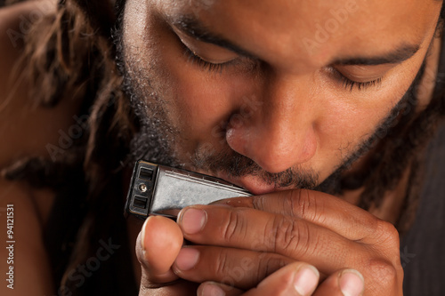 Fotografie A close-up of an Afro man with dreadlocks and a beard playing a diatonic harmonica with his eyes closed in focus, warm soft lighting highlighting the soulful intimate mood of the blues music