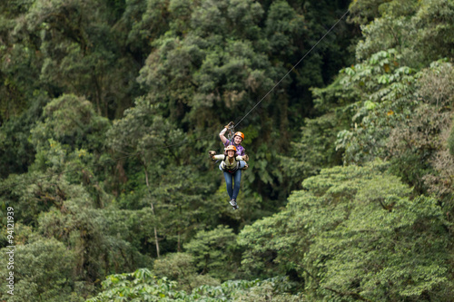 A woman wearing sports gear slides down a zipline through the lush jungle canopy, holding onto a rope as she speeds along the line.