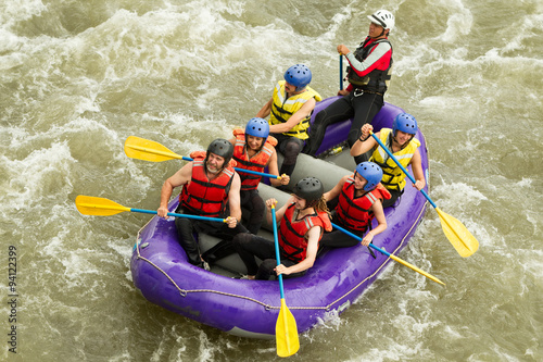 A family of four smiling on a white water raft, navigating through choppy waters with splashes of white foam surrounding them.