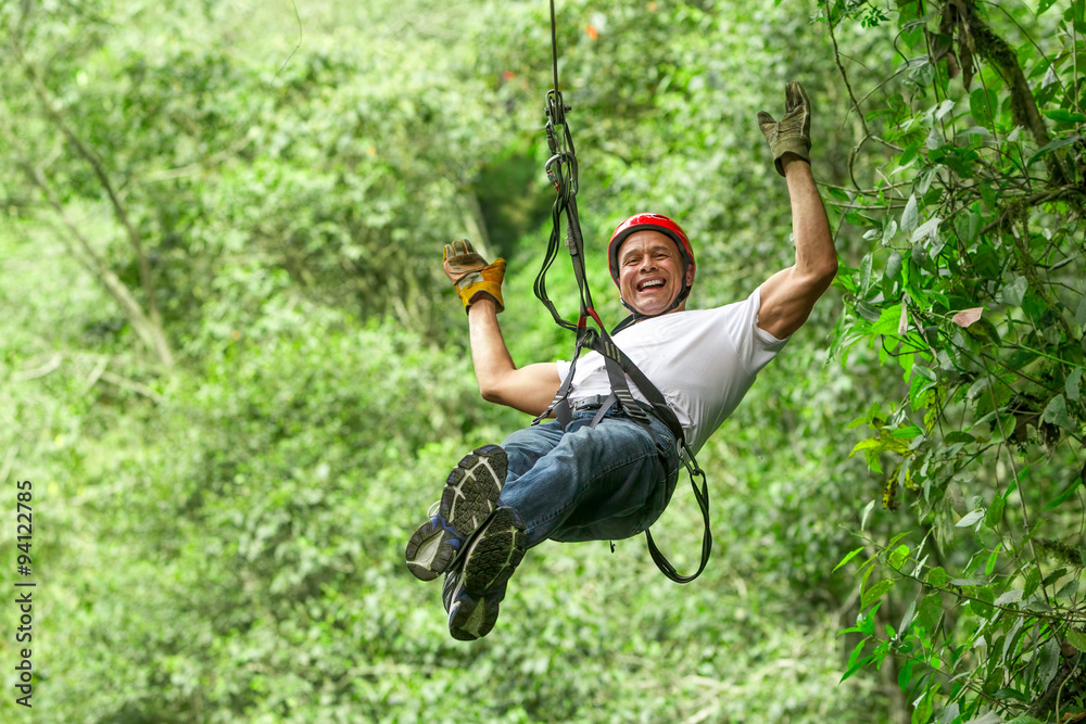 An adult man wearing a harness is ziplining through the forest, feeling the adrenaline rush as he speeds along a rope line.