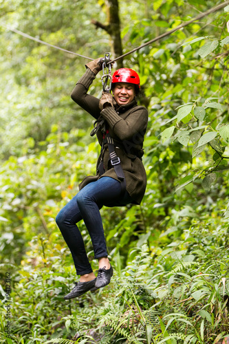 A black-clad woman fearlessly climbs a zipline, suspended on a wire, surrounded by other people enjoying the thrilling adventure.