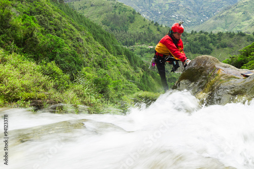 A canyoning guide in a red jacket and helmet steps across rocks in a fast-flowing river amid green mountains at Chamana near Banos, Ecuador, a dynamic outdoor adventure.