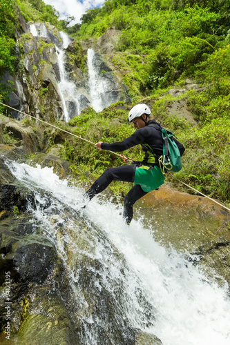 A daring adventurer rappels down a steep cliff in the lush canyons of Baños, Ecuador, with a stunning waterfall cascading beside them.