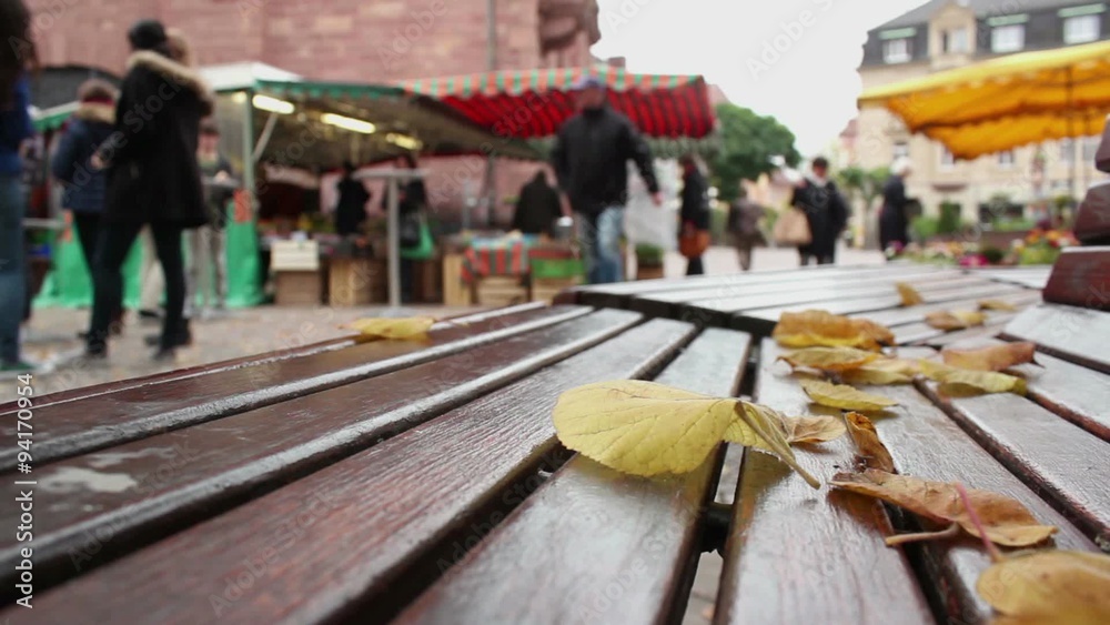 Green market at central city square (marktplatz) in Ettlingen - Baden Wurttemberg, Germany; few autumn leaves at park bench with blurry people in background; Green market at central city square (marktplatz) in Ettlingen - Baden Wurttemberg, Germany; few autumn leaves at park bench with blurry people in background;