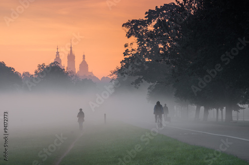 Tree alley along Blonia meadow in Krakow  Poland  with St Mary s church and Town Hall towers in the background  foggy morning with people s silhouettes