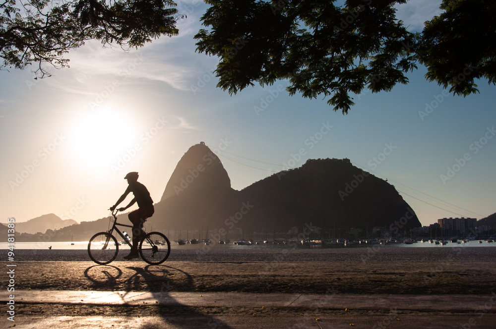 Naklejka premium Silhouette of a Man Cycling in the Early Morning during Beautiful Warm Sunrise in Rio de Janeiro