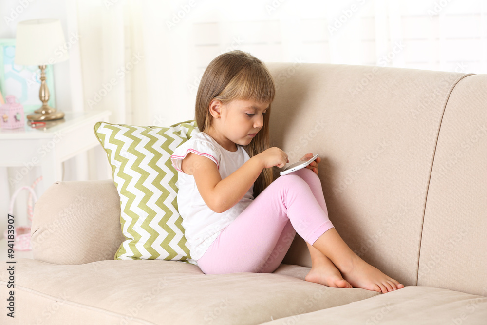 Beautiful little girl  with smart phone sitting on sofa, on home interior background