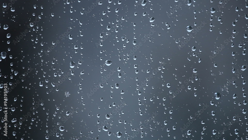 Water drops from melted snow and rare snowflakes on window glass against grey background. Snowing, stormy weather, movement of snowflakes visible through glass.
