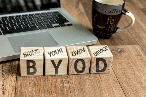 Bring Your Own Device (BYOD) written on a wooden cube in front of a laptop photo