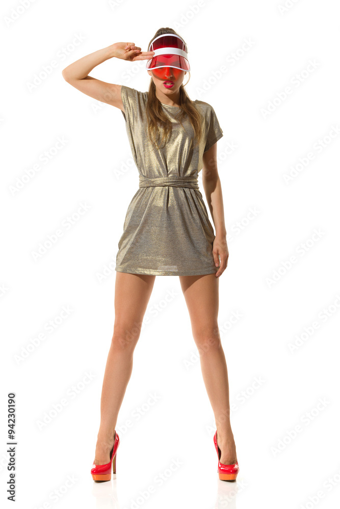 Serious fashion young woman in gold mini dress, red high heels and sun visor cap standing with legs apart and salute. Full length studio shot isolated on white. Serious fashion young woman in gold mini dress, red high heels and sun visor cap standing with legs apart and salute. Full length studio shot isolated on white.