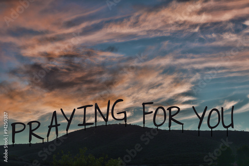 Praying for you. The words silhouetted as on a hillside with dramatic pink clouds and sky.
