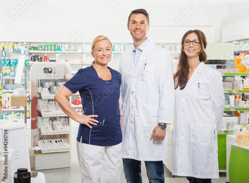 Female Assistant With Pharmacists Standing In Pharmacy