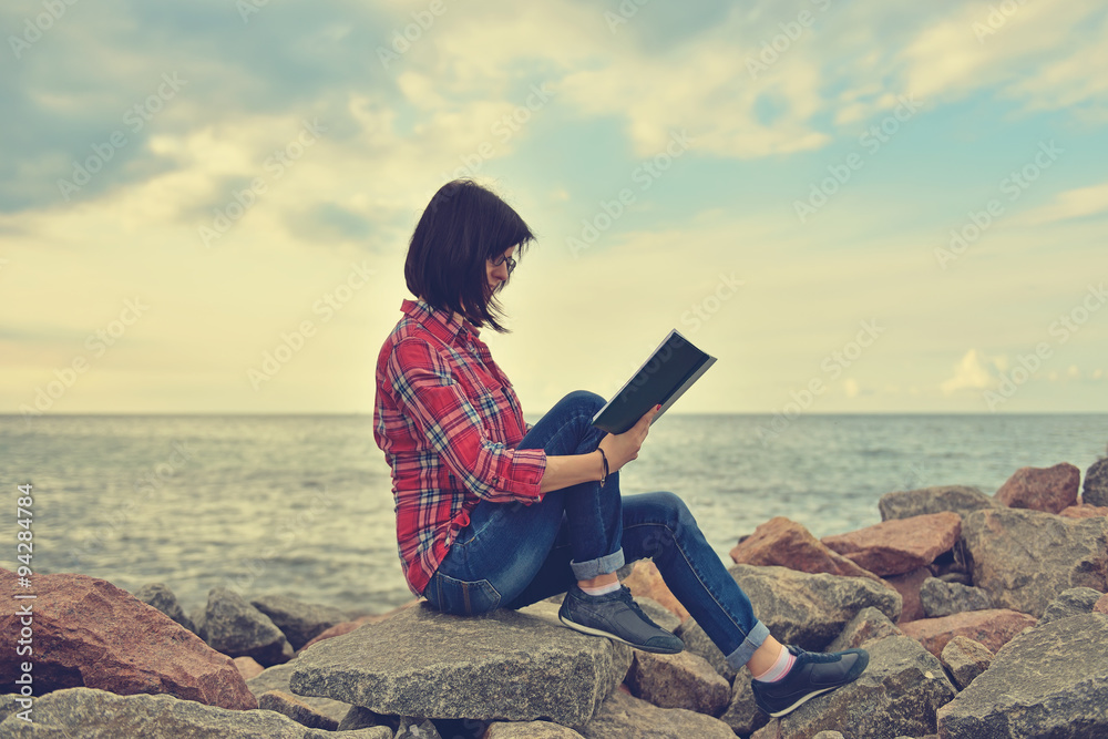 young hipster girl sitting on rock with book
