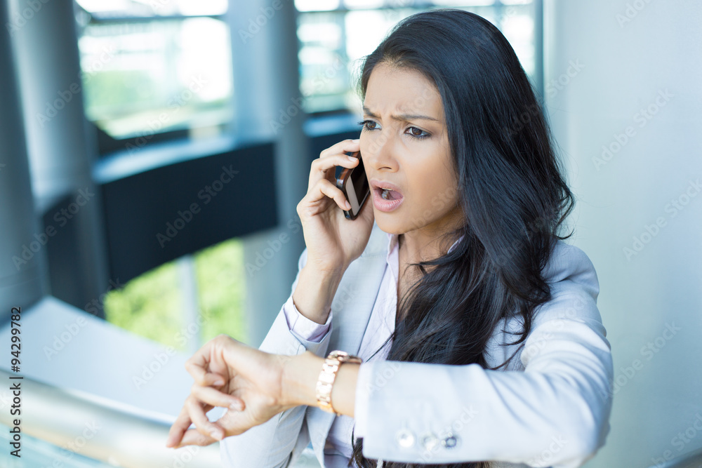 Closeup portrait, young woman in gray business suit blazer talking on cell phone concerned about running out of time on watch, isolated indoors office background Closeup portrait, young woman in gray business suit blazer talking on cell phone concerned about running out of time on watch, isolated indoors office background